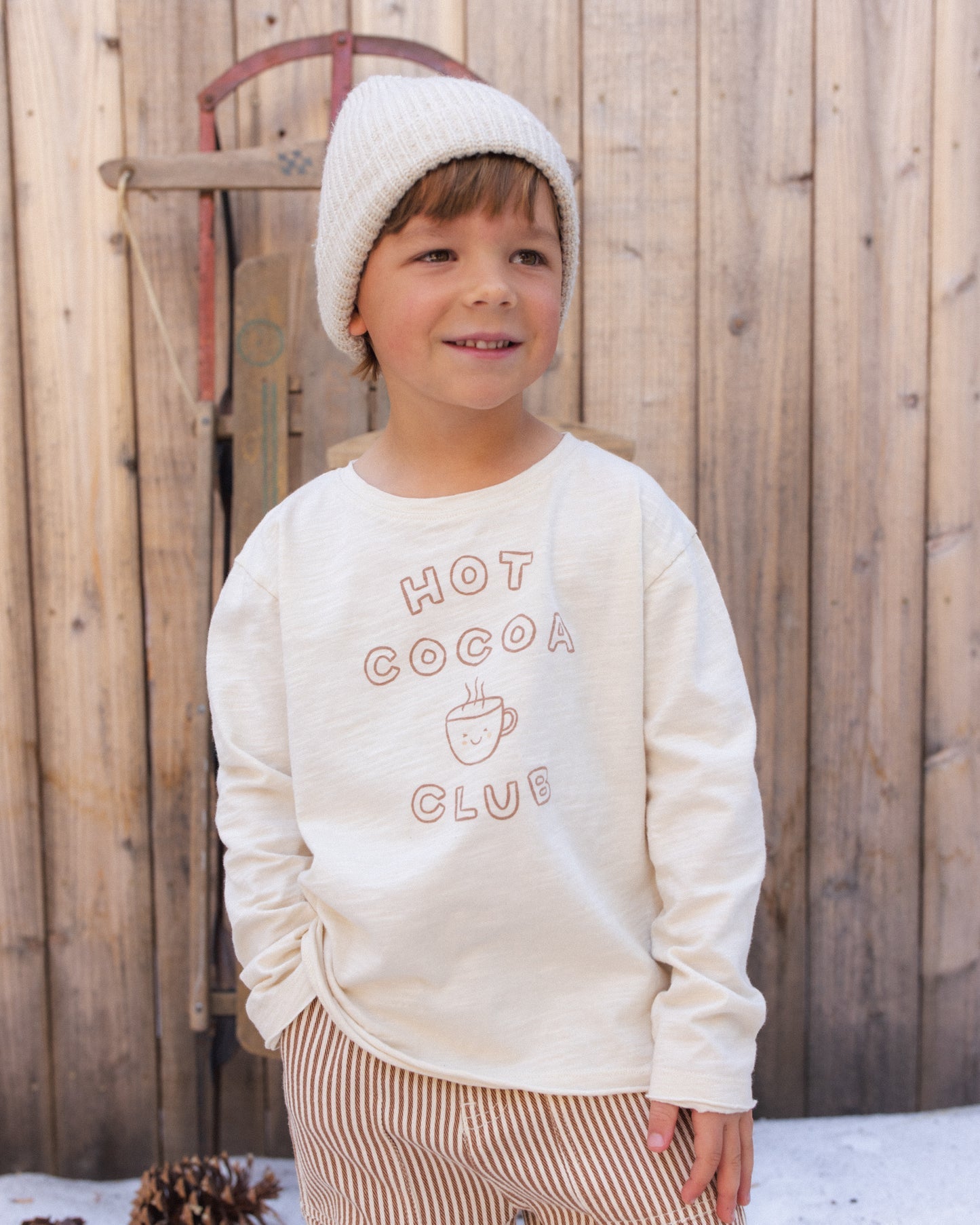 A young child wearing a cream-colored long-sleeve sweatshirt with 'HOT COCOA CLUB' printed on the front, paired with a matching knit beanie and striped pants, standing in front of a rustic wooden fence.