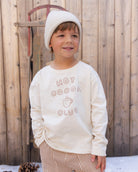 A young child wearing a cream-colored long-sleeve sweatshirt with 'HOT COCOA CLUB' printed on the front, paired with a matching knit beanie and striped pants, standing in front of a rustic wooden fence.