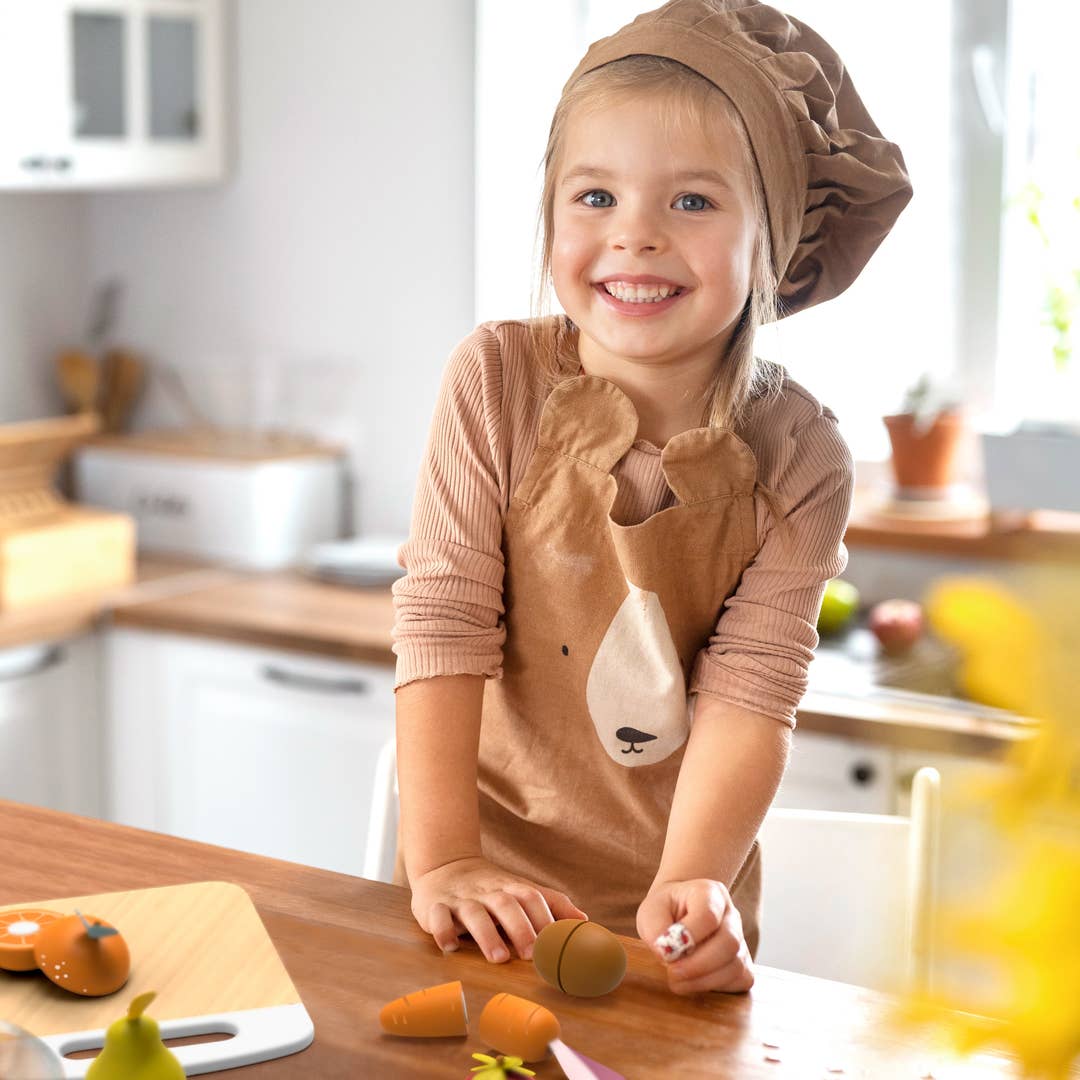 A young child wearing a brown chef hat and a matching brown apron with a bear face design stands at a wooden table in a bright kitchen, holding toy vegetables.