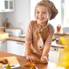 A young child wearing a brown chef hat and a matching brown apron with a bear face design stands at a wooden table in a bright kitchen, holding toy vegetables.
