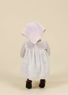 Child wearing a white dress with lace details and a pink hood on a beige background