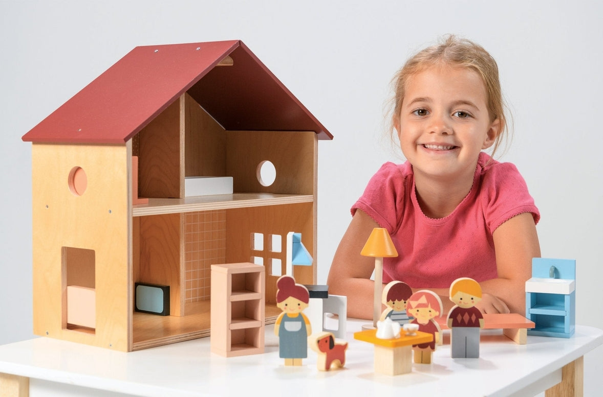 A young child playing with an open-faced wooden dollhouse by Mentari, arranged on a white table with miniature wooden furniture and small figurines.