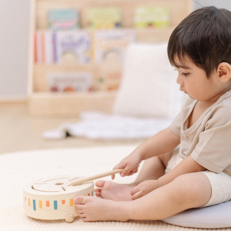 A young child sitting on the floor plays with a wooden Plan Toys sensory drum using a small mallet.