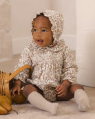 A baby wearing a cream and brown floral smocked romper with a matching ruffled bonnet, sitting on a carpeted floor next to a wicker basket.