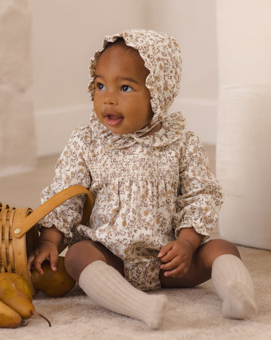A baby wearing a cream and brown floral smocked romper with a matching ruffled bonnet, sitting on a carpeted floor next to a wicker basket.
