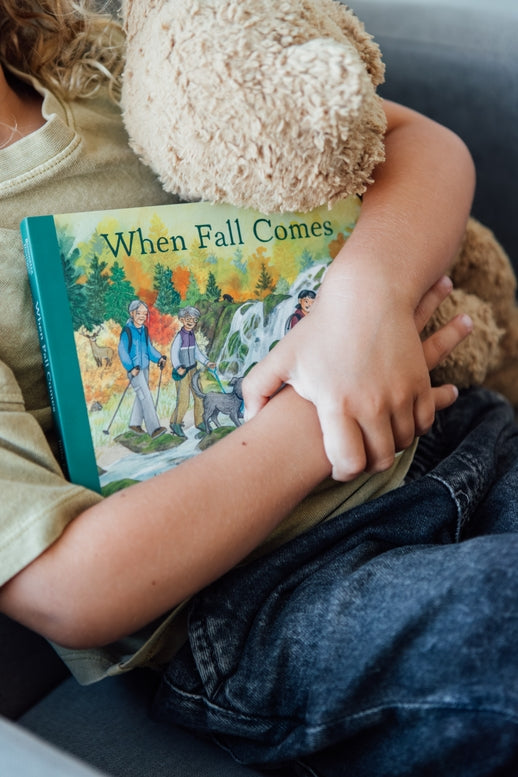 A toddler holding an open board book titled When Fall Comes, looking down at the colorful illustrated pages showing autumn trees and an outdoor nature scene.