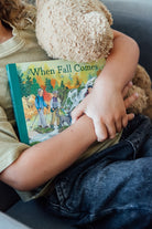 A toddler holding an open board book titled When Fall Comes, looking down at the colorful illustrated pages showing autumn trees and an outdoor nature scene.