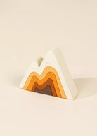 A minimalist mountain-shaped decorative object with layered concentric wave patterns in white, light beige, orange, brown, and dark brown, set against a solid cream background.