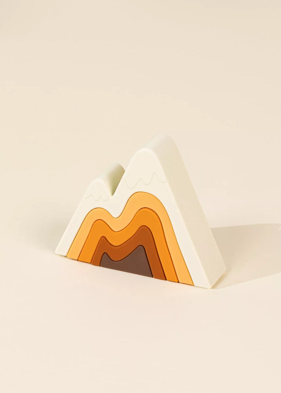 A minimalist mountain-shaped decorative object with layered concentric wave patterns in white, light beige, orange, brown, and dark brown, set against a solid cream background.