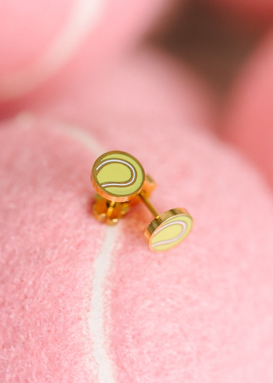 Tennis ball-themed stud earrings on a pink fabric background