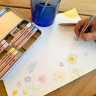 Person coloring with pastel-colored pencils on a piece of paper with floral designs, next to a blue glass and a metal case of pencils.
