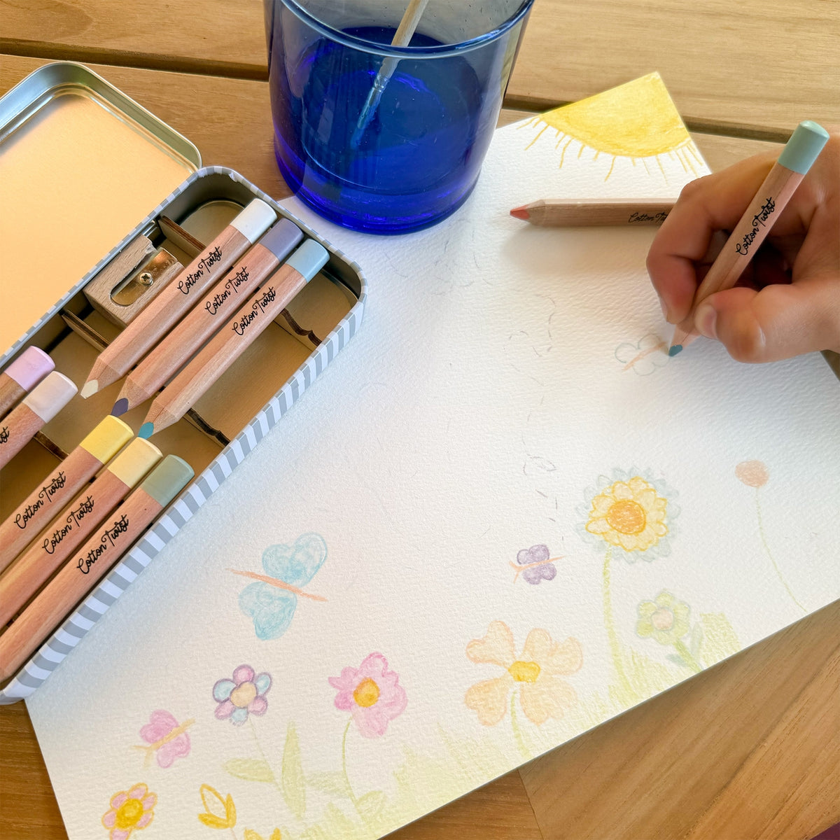 Person coloring with pastel-colored pencils on a piece of paper with floral designs, next to a blue glass and a metal case of pencils.