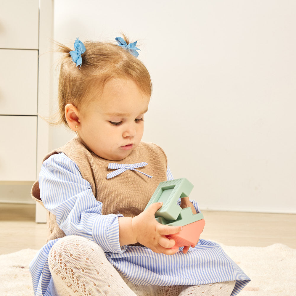 A toddler girl sitting on a light-colored rug, holding a pastel-colored wooden toy. She is wearing a blue and white striped dress, a brown vest, and has two blue bow hair clips.
