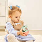 A toddler girl sitting on a light-colored rug, holding a pastel-colored wooden toy. She is wearing a blue and white striped dress, a brown vest, and has two blue bow hair clips.
