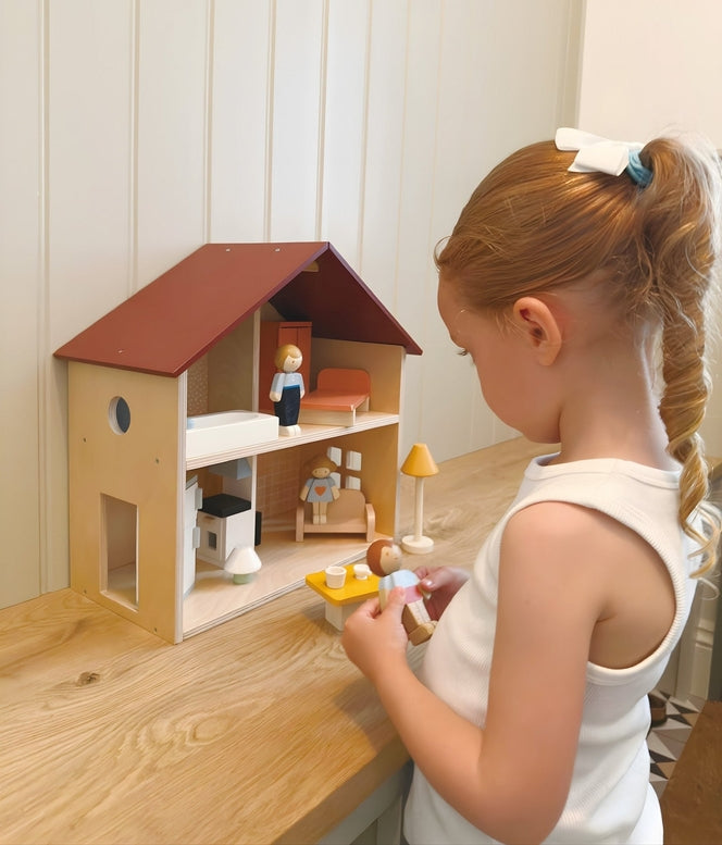 A young child playing with an open-faced wooden dollhouse by Mentari, arranged on a white table with miniature wooden furniture and small figurines.
