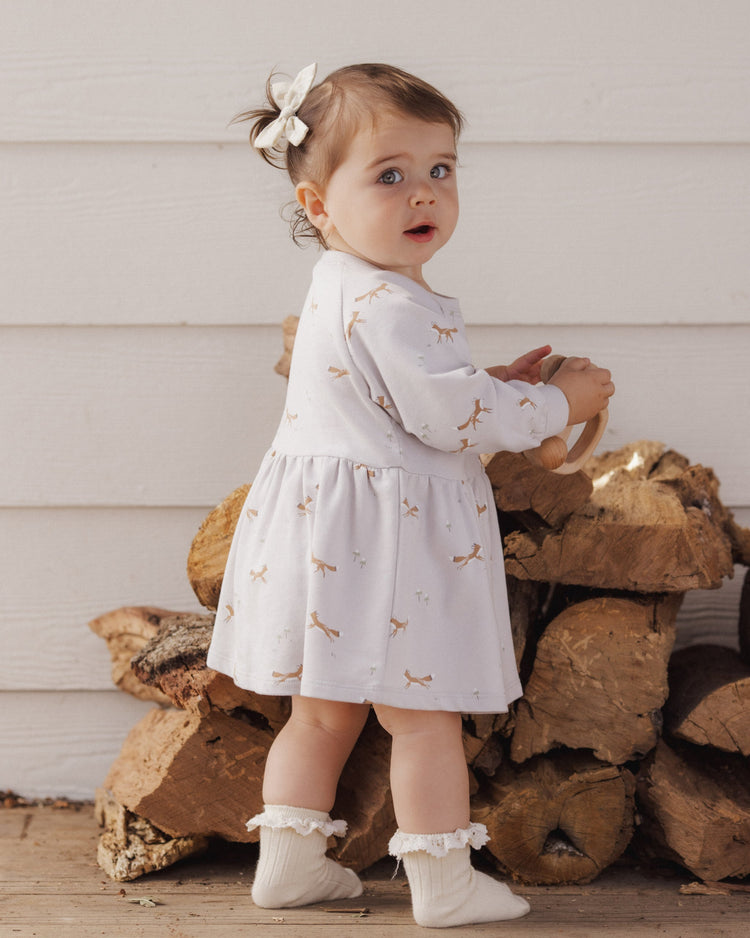 A toddler wearing the Rylee + Cru Foxes Raglan Dress in a soft fog hue, styled with a sweet matching bow and ruffled socks.