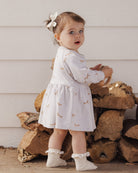 A toddler wearing the Rylee + Cru Foxes Raglan Dress in a soft fog hue, styled with a sweet matching bow and ruffled socks.