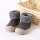 Pair of gray baby shoes with beige soles next to a woven basket on a white background