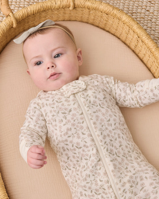 A baby lying in a woven bassinet wearing a cream-colored long-sleeve footless sleeper with a delicate floral pattern and a matching bow headband.