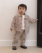 A toddler boy wearing a matching two-piece pajama set with brown and beige vertical stripes, consisting of a long-sleeve button-up shirt and pants, standing against a white wall.