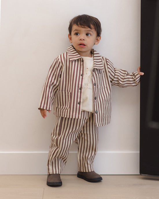 A toddler boy wearing a matching two-piece pajama set with brown and beige vertical stripes, consisting of a long-sleeve button-up shirt and pants, standing against a white wall.