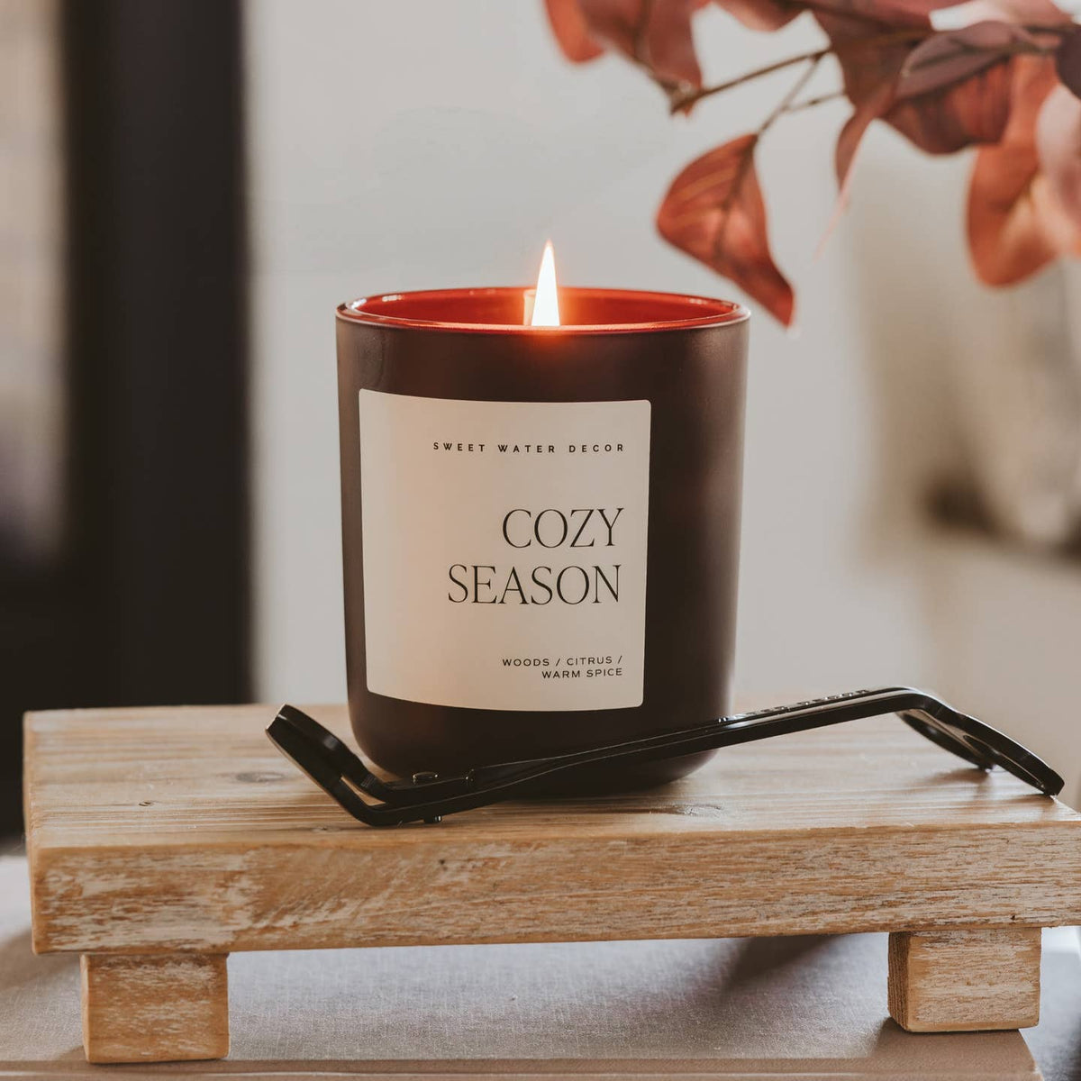 Candle labeled 'Cozy Season' on a wooden stand with blurred background