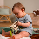 A baby sitting on a play mat, holding a soft fabric sensory toy with a green handle. The baby is wearing a light blue muslin romper in a softly lit room with a wicker chair and basket in the background.