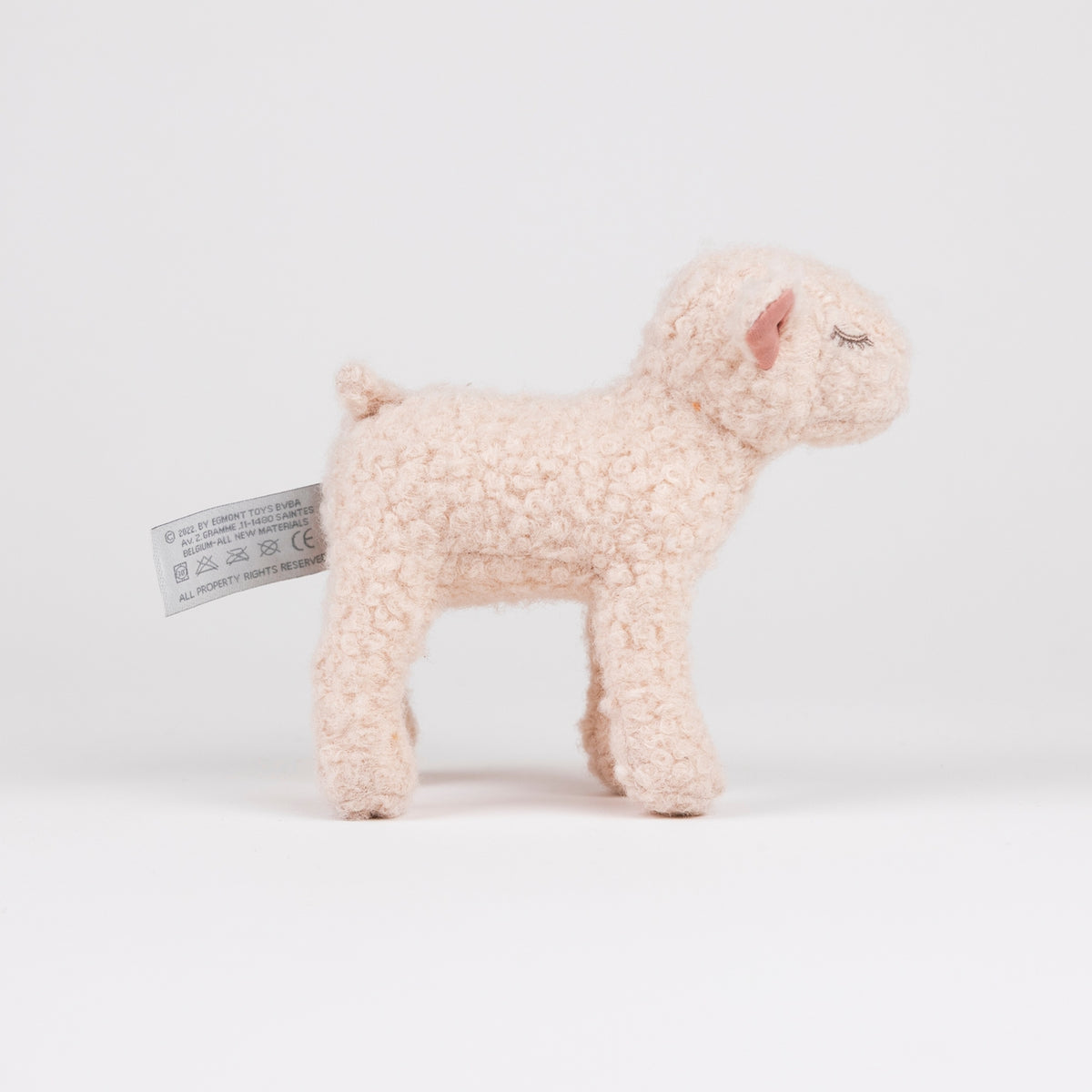 A small plush toy lamb made from curly, textured yarn in a pale peach-pink color, standing on a seamless white studio background. A white product tag is attached to its side.
