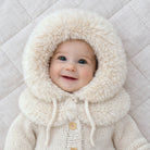 Baby wearing a cream-colored fluffy hooded outfit on a light gray quilted background