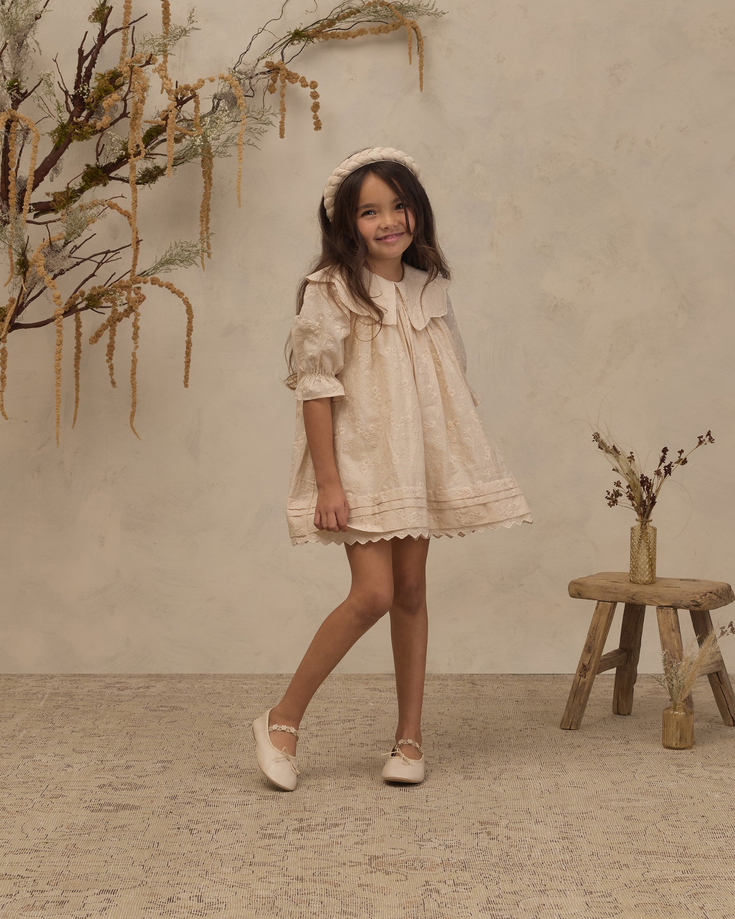 A young girl wearing a cream-colored lace dress with a matching headband and white shoes, standing in a styled studio setting with a textured beige wall, dried floral decorations, and a small wooden stool.