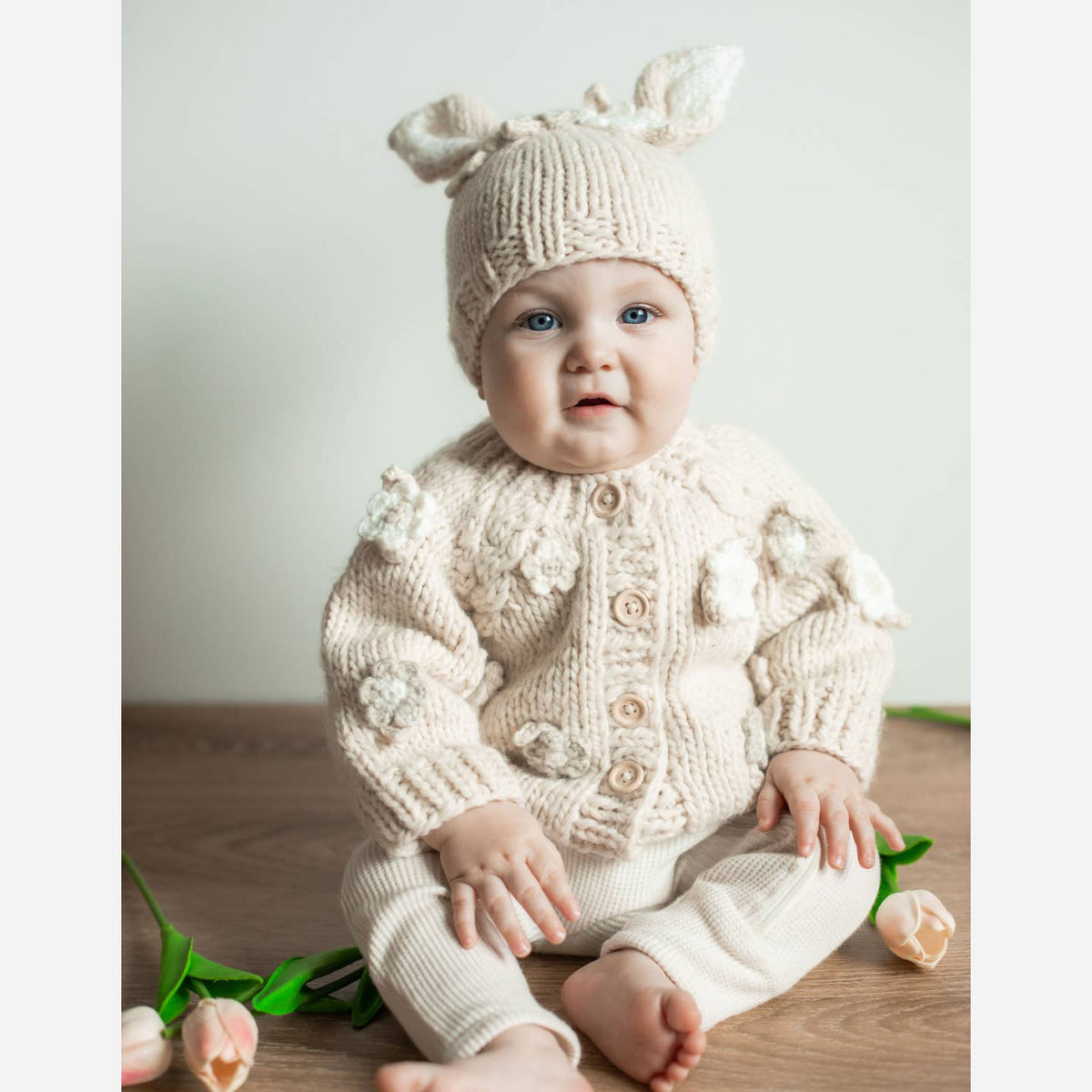 Baby wearing a cream-colored knitted outfit with a matching hat, sitting on a wooden floor.