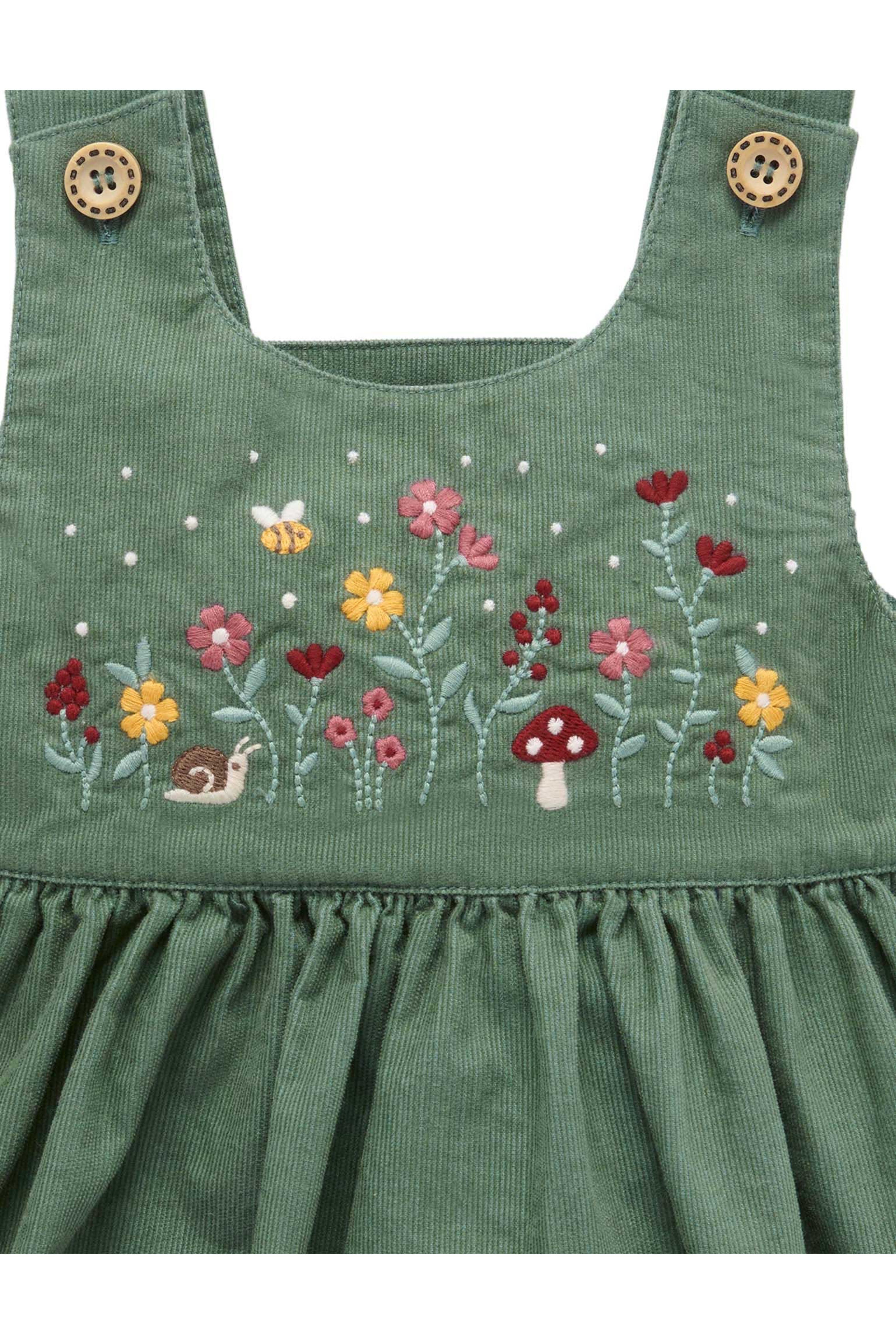 Green corduroy baby dress featuring colorful embroidered wildflowers, a mushroom, a bee, and a snail on the chest, with decorative wooden buttons on the shoulder straps.