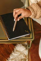 Person holding a pen over a black notebook with gold spiral binding on a gold tray.