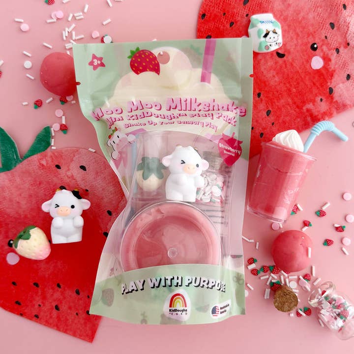 A toy milkshake set in a clear bag with a pastel green label reading 'Moo Moo Milkshake' and 'Play with Purpose.' The package contains a pink drink cup, a small white cow figure, and a strawberry-themed accessory, arranged on a pink surface with watermelon and candy props.