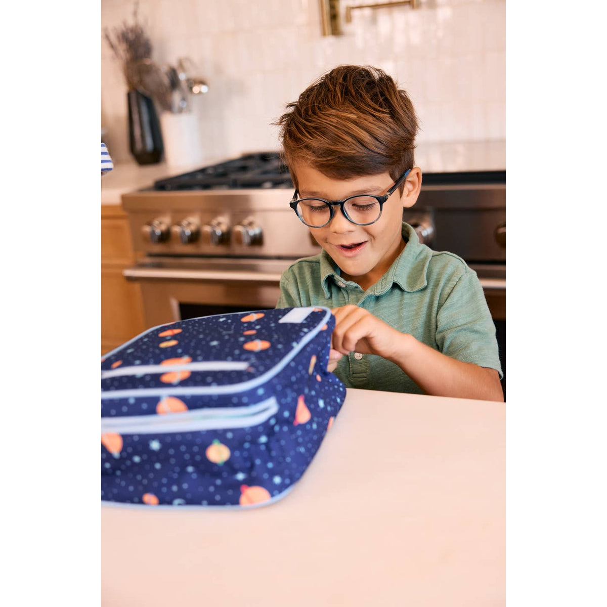 Child with glasses opening a blue lunch bag with pink hearts in a kitchen.