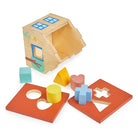 A wooden house shape sorter with its red roof panel detached and laid flat, revealing the interior while five colorful wooden shapes—a mint green cube, yellow cylinder and triangle, pink heart, and light blue cross—sit in front of the toy.