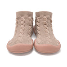 A pair of slip-on knit socks with a braided cable pattern in beige and a dusty pink rubber sole, shown from the front on a white background.