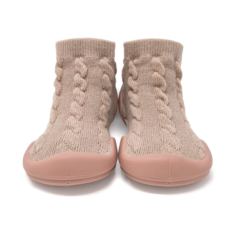 A pair of slip-on knit socks with a braided cable pattern in beige and a dusty pink rubber sole, shown from the front on a white background.