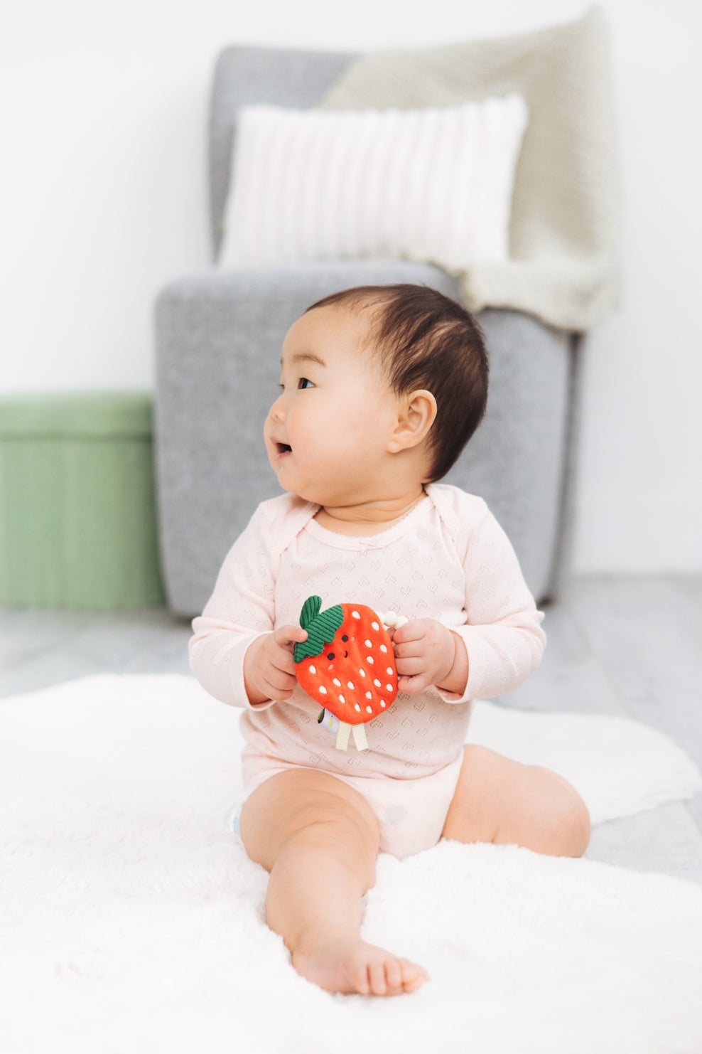 Baby holding a strawberry-shaped toy on a white blanket with a gray cushion in the background