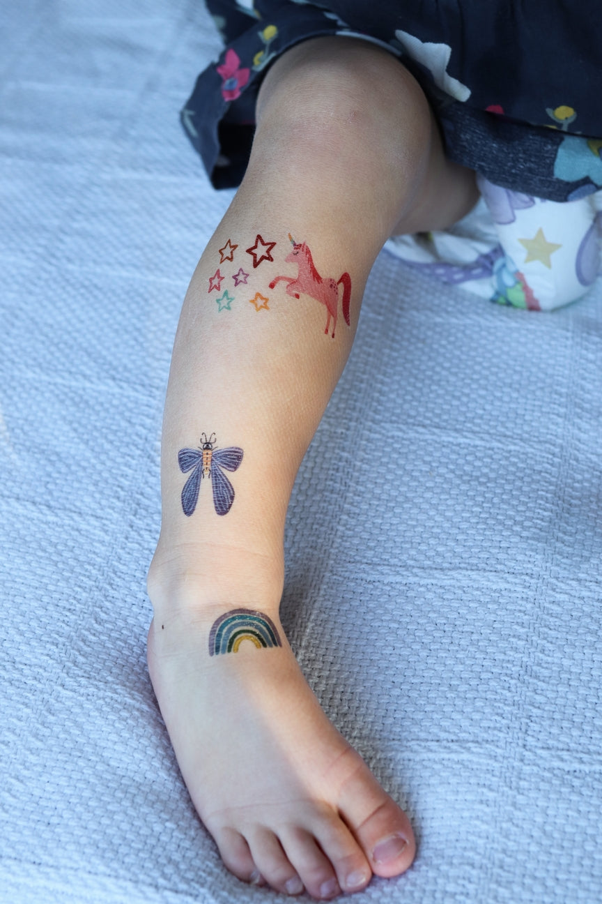 Child's leg with temporary tattoos of a unicorn, rainbow, and butterfly on a textured surface.