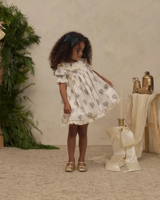 A young girl with curly hair wearing a cream-colored dress with a gray floral pattern and a teal bow headband, standing in a styled studio setting with a beige textured wall, greenery, and a table with a large cream ribbon.