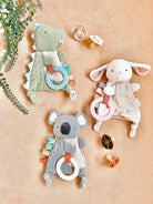 Three soft animal toys with rings on a beige surface, surrounded by leaves and pacifiers.