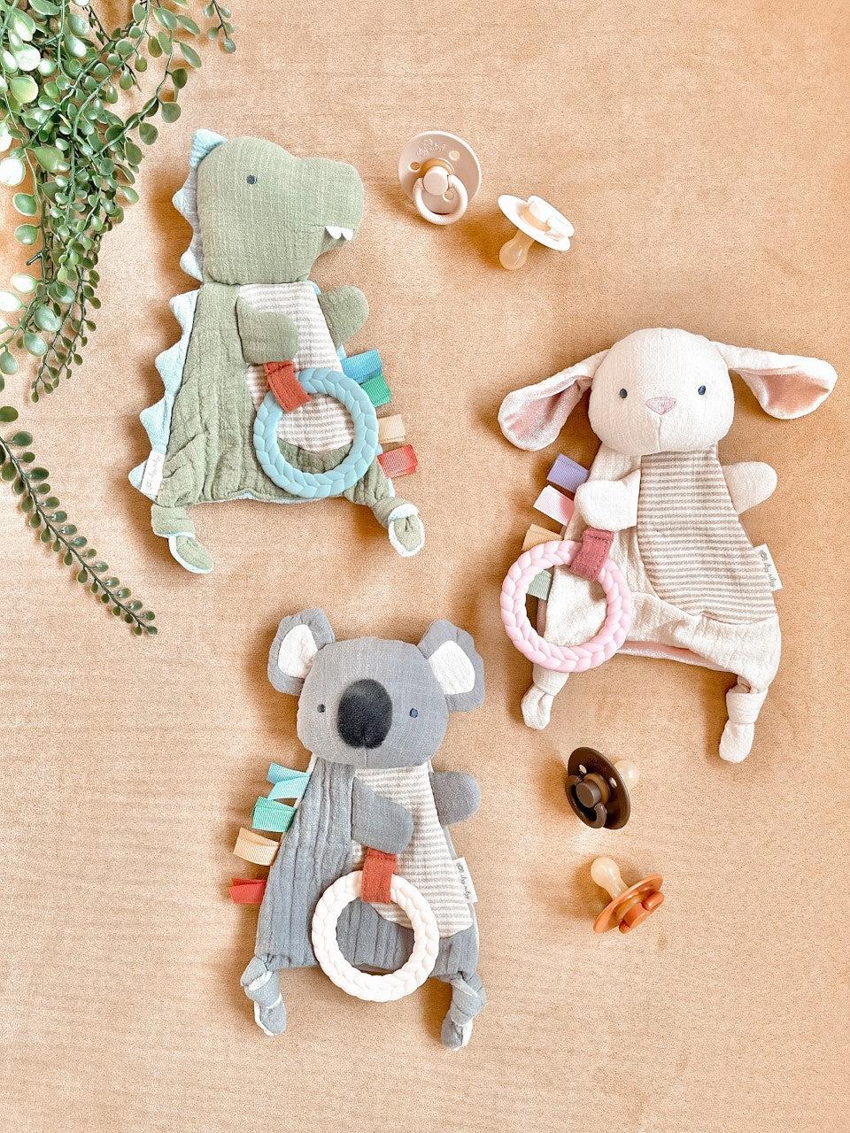 Three soft animal toys with rings on a beige surface, surrounded by leaves and pacifiers.