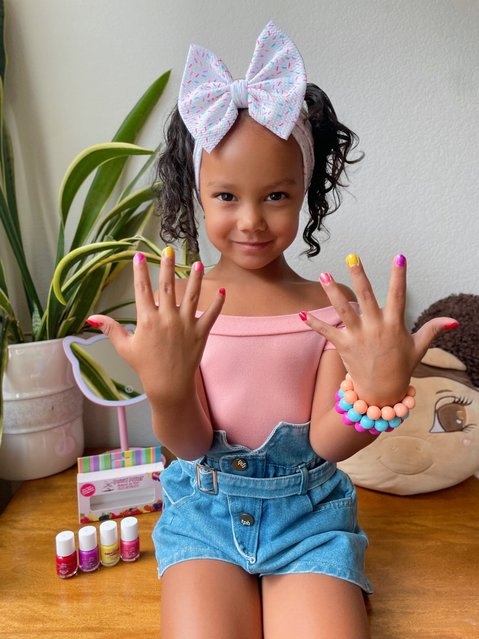 Young girl showing off her painted nails with a headband and colorful bracelet, surrounded by nail polish bottles and a plant.
