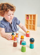 A toddler with curly hair sits at a light wood table playing with colorful wooden peg dolls, with a small wooden storage box in the background.