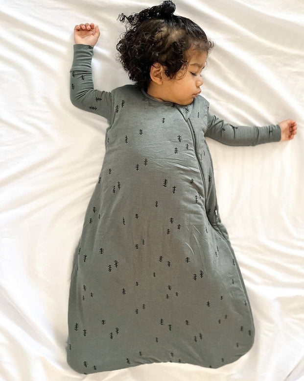 A baby wearing a gray long-sleeve sleeping bag with a small black tree pattern, lying on white bedding.