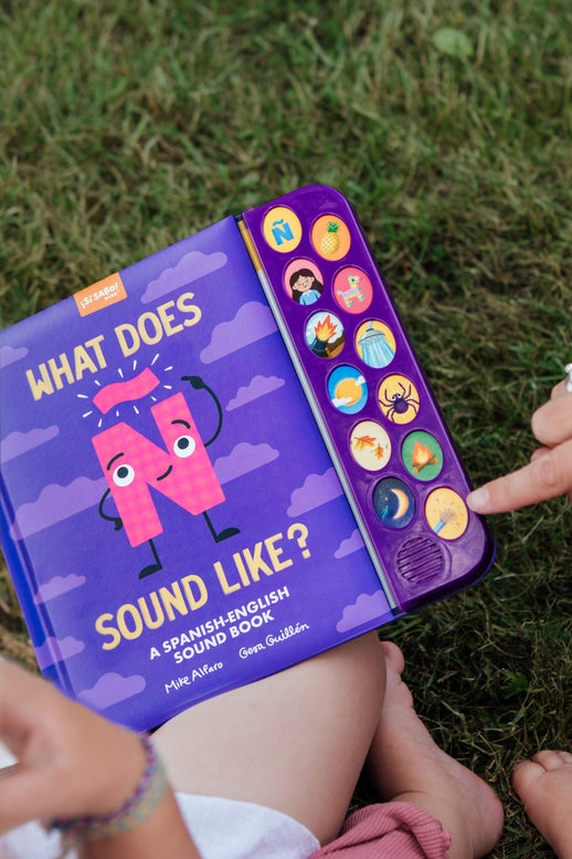 A person holding "What Does Ñ Sound Like?: A Spanish-English Sound Book" outdoors on grass, showing the purple cover with an animated letter Ñ character, clouds, and a vertical strip of interactive sound buttons.