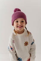 A young child wearing a pink ribbed knit pom-pom beanie and a cream knit sweater with colorful embroidered flowers, photographed against a plain light background.