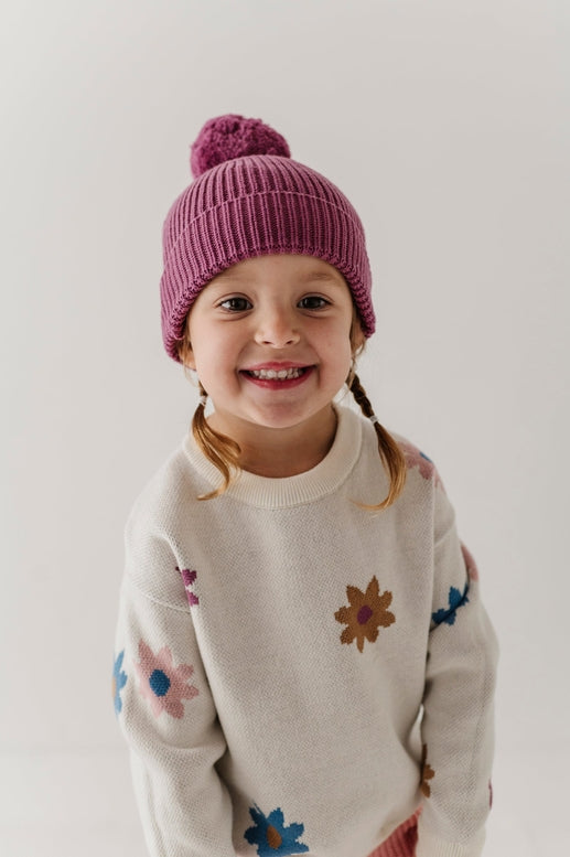 A young child wearing a pink ribbed knit pom-pom beanie and a cream knit sweater with colorful embroidered flowers, photographed against a plain light background.