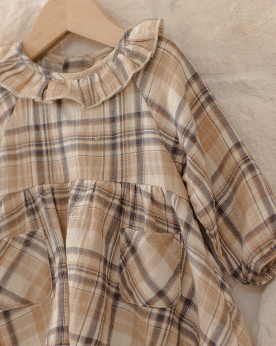 A close-up of a children's plaid dress in beige, brown, and gray tones, hanging on a wooden hanger against a textured beige fabric background.
